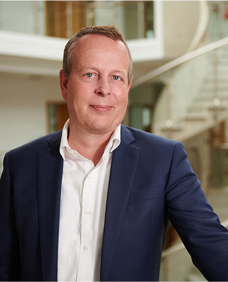 Geschäftsmann in Anzug lächelt vor einer modernen Treppe in einem hellen Raum.