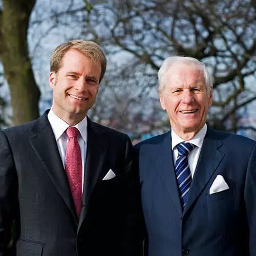Zwei Männer in Anzügen lächeln vor einem Baum, symbolisieren Geschäft und Partnerschaft.