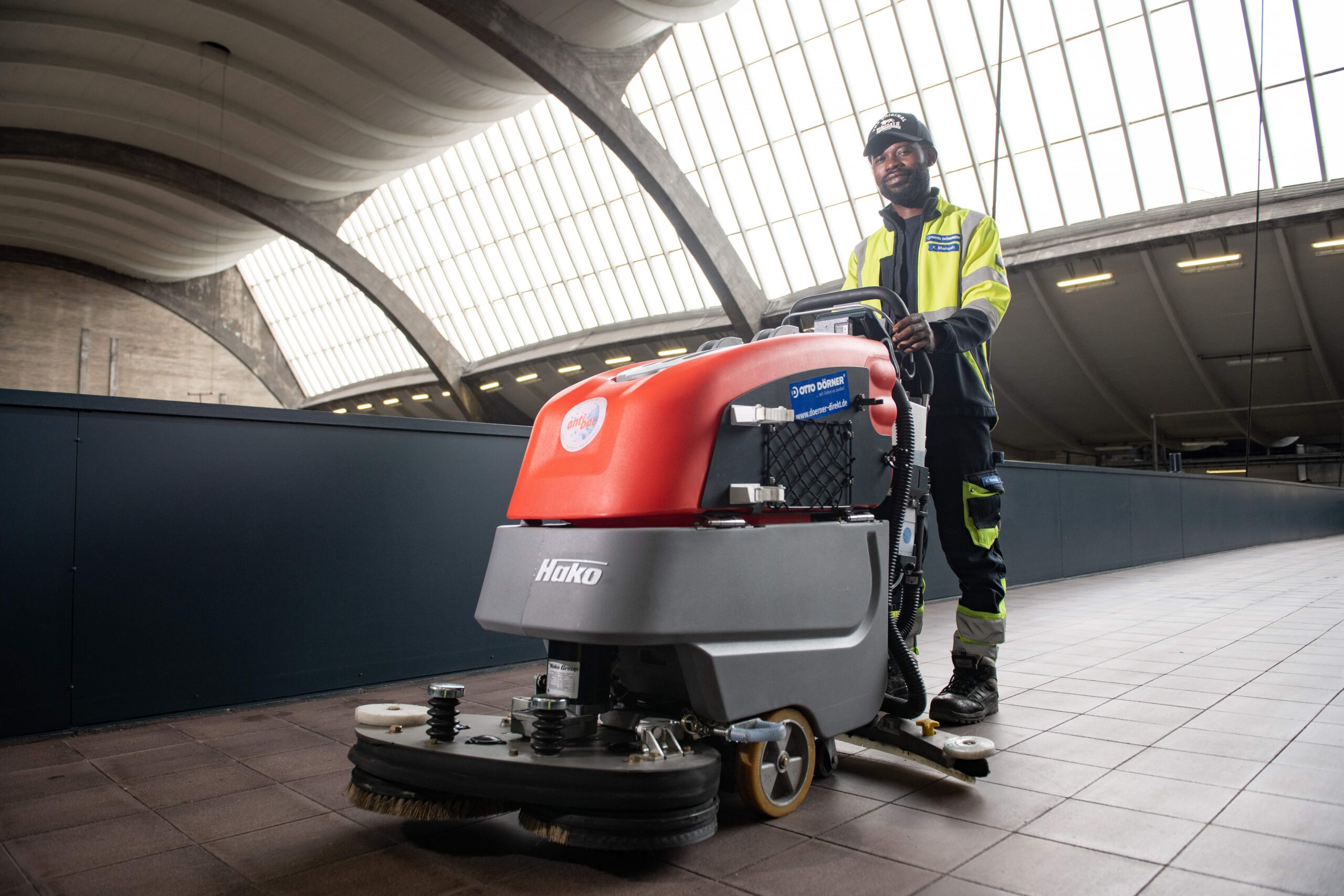 Mitarbeiter mit Bodenreinigungsmaschine in modernem Geb&auml;ude mit gro&szlig;en Fenstern.